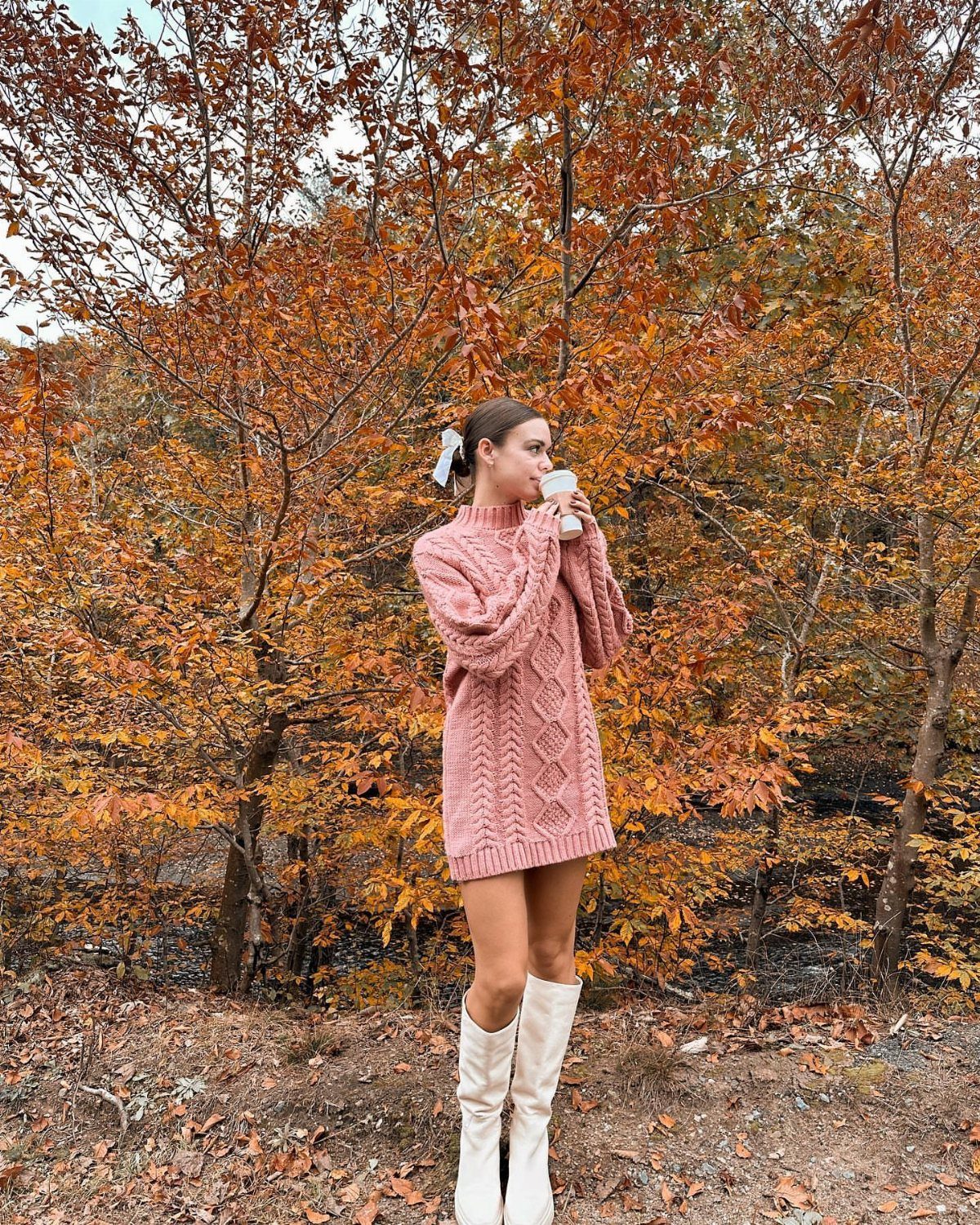 Woman in a pink oversized cable-knit sweater dress and white boots standing among autumn trees.