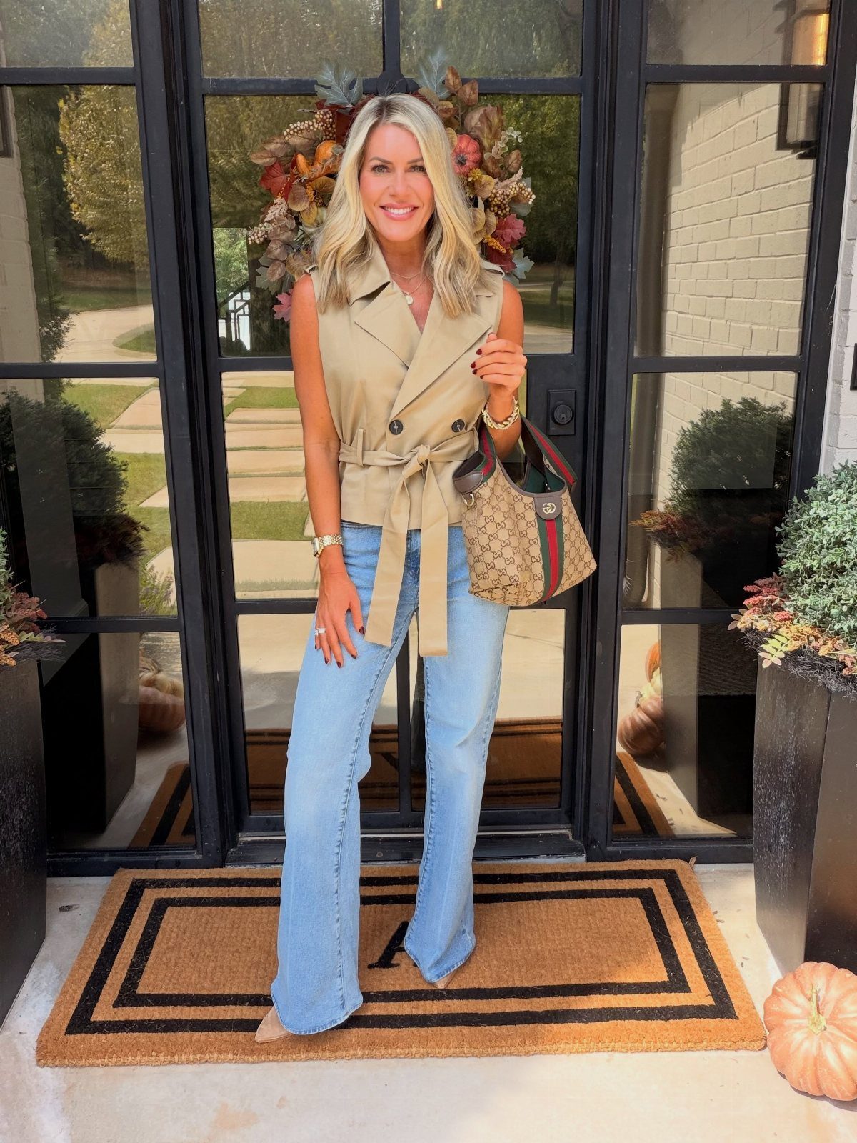 Smiling woman wearing a tan tailored vest and wide-leg blue jeans standing by a glass door.