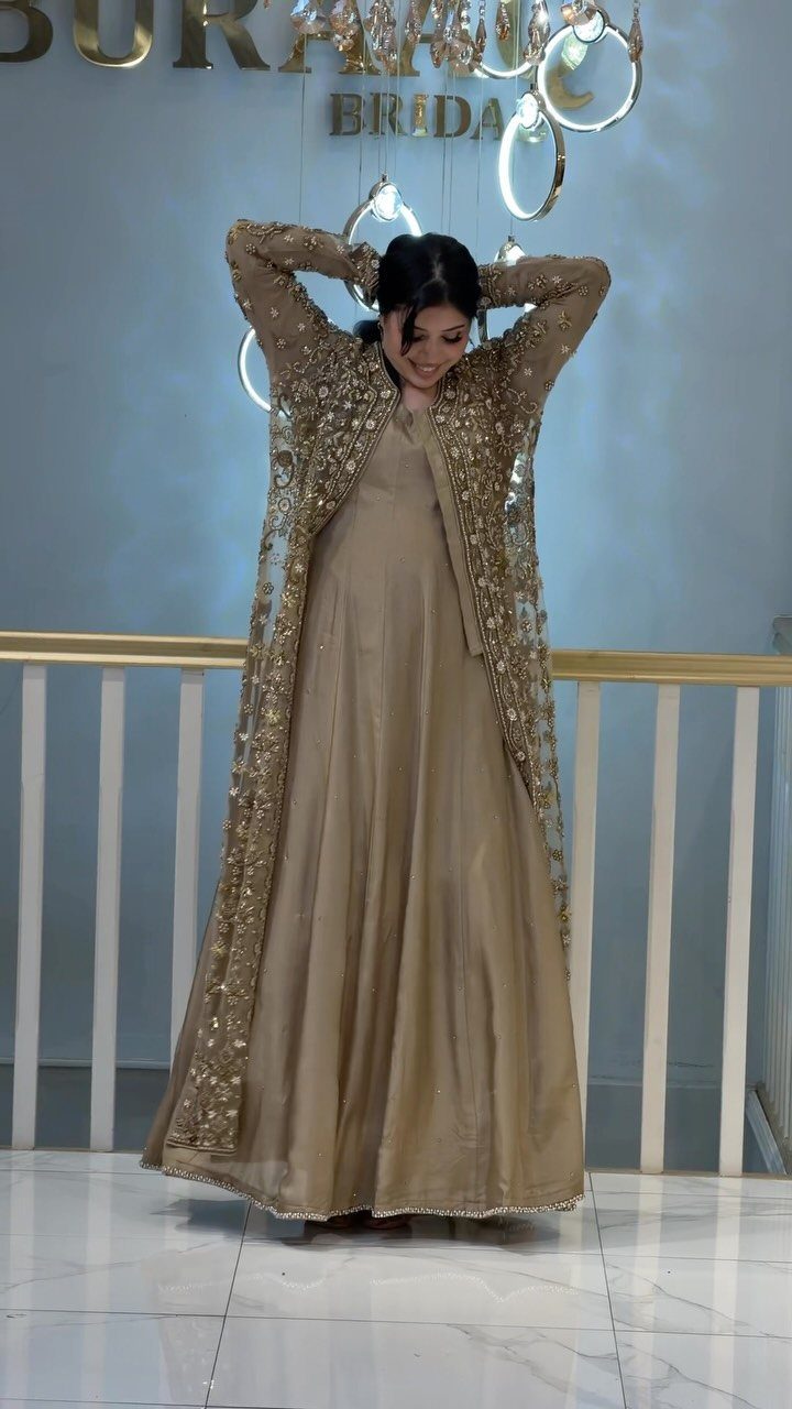 Woman in a champagne satin gown and gold embroidered lace jacket standing indoors.
