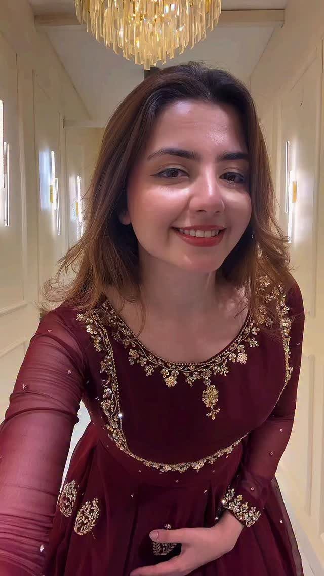 A smiling woman wearing a maroon dress with intricate gold embroidery and long sheer sleeves.