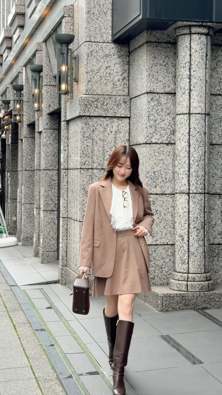 Woman in a tan blazer and matching skirt walking past a stone building with columns.