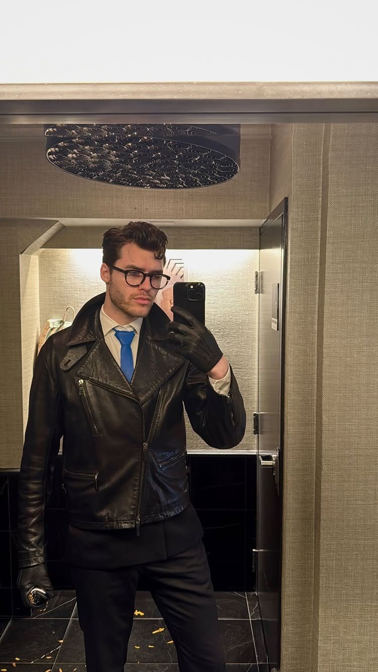 Man in a black leather jacket, white button-down shirt, blue tie, and dark trousers taking a selfie.
