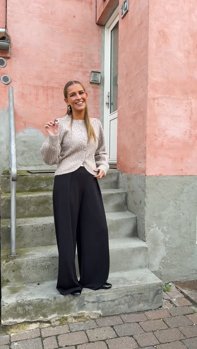 Smiling woman wearing a beige knit cardigan and black wide-leg trousers standing on outdoor stairs.