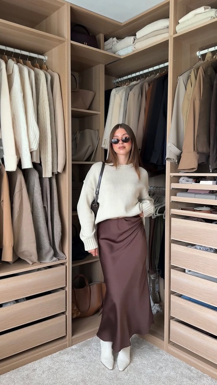 Woman in a cream sweater, brown satin skirt, and white boots standing in a closet.