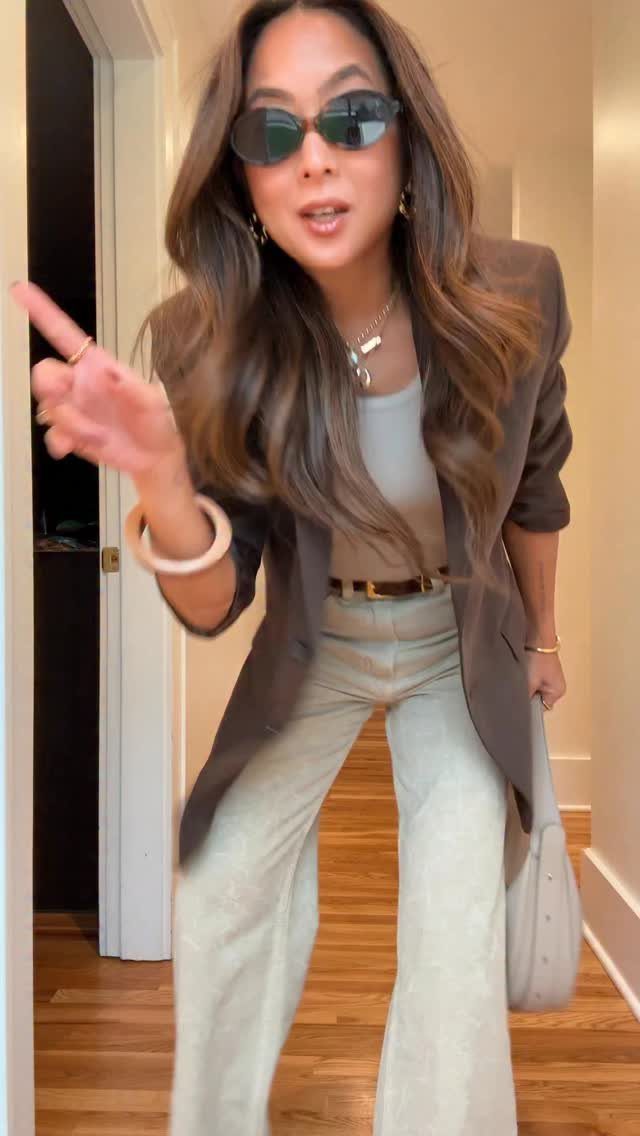 Woman in a brown blazer and light wide-leg trousers posing in a hallway.