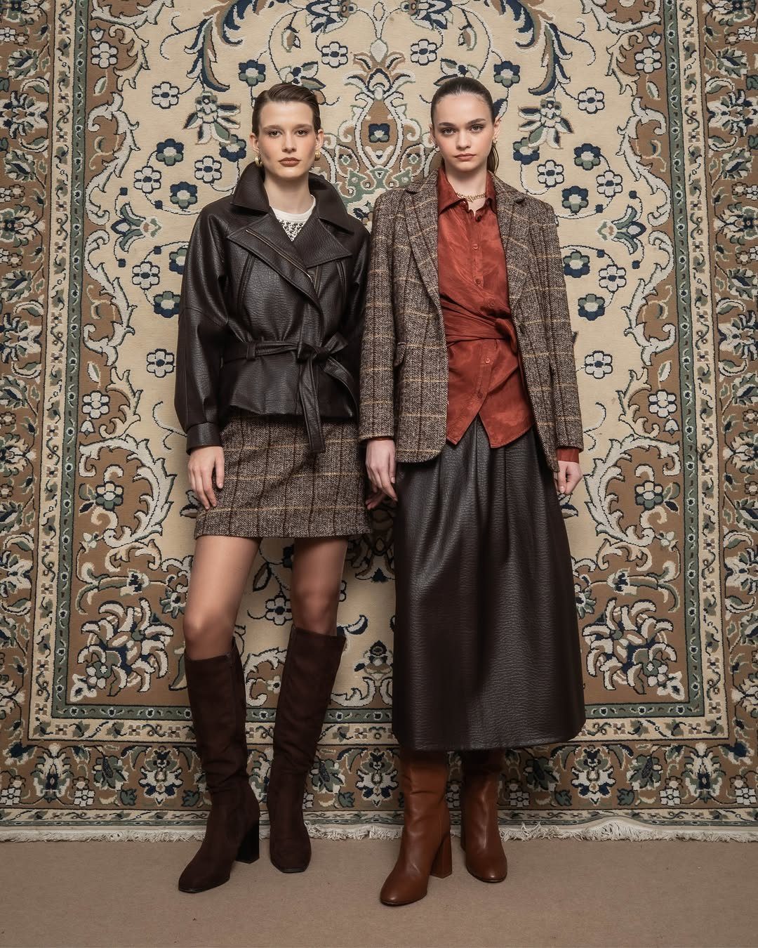 Two women posing in professional autumn outfits against a large patterned tapestry background.