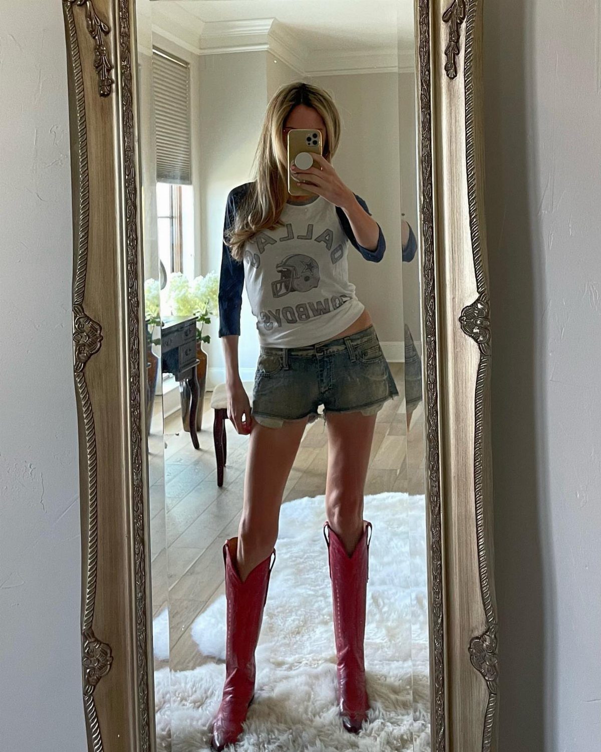 Woman posing in a mirror wearing a graphic tee, denim shorts, and bright pink cowboy boots.