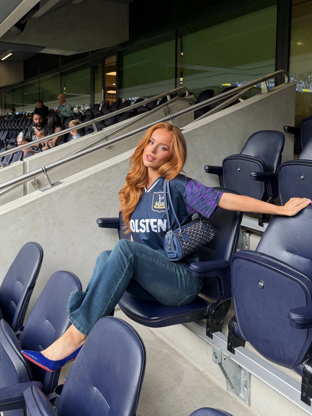Red-haired woman sitting in stadium seats wearing a blue football jersey and denim jeans.