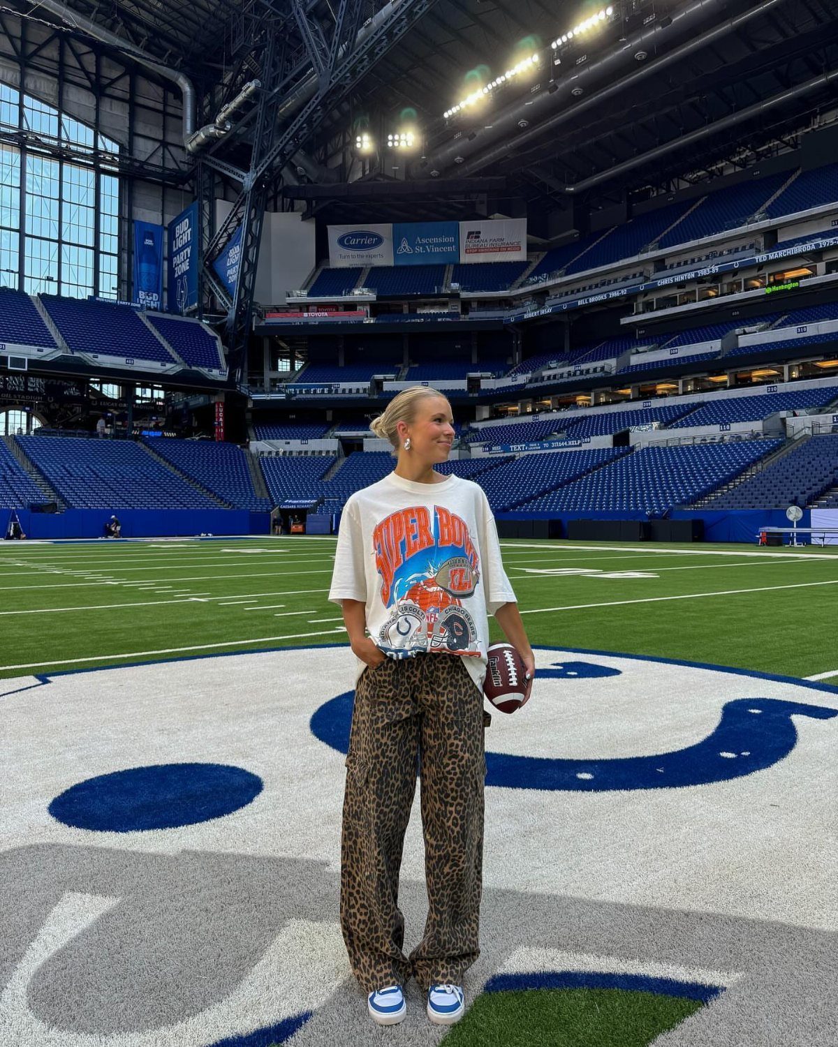 Woman in an oversized graphic tee and leopard print cargo pants standing on a football field.