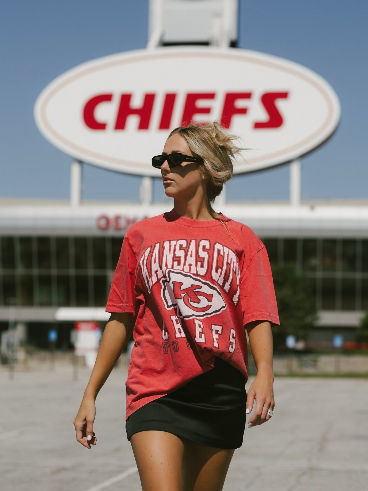 Woman in a red Kansas City Chiefs t-shirt and black skirt standing before a stadium sign.