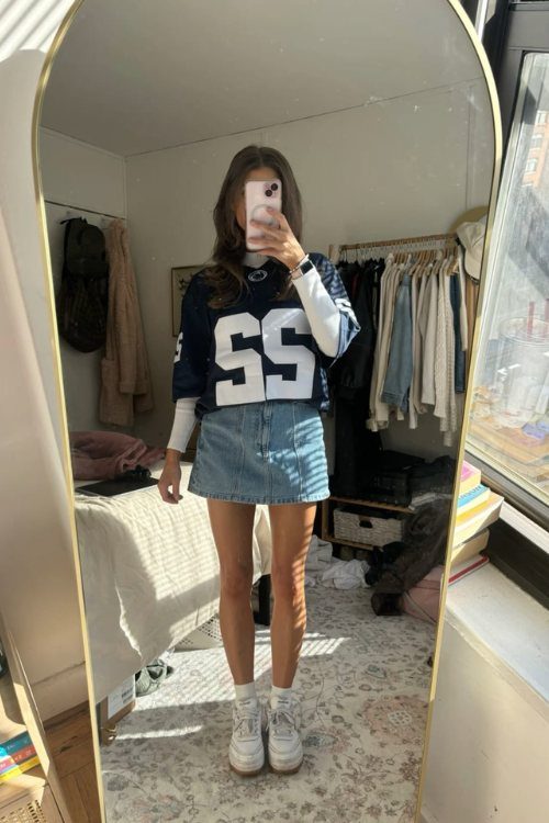 Woman in a navy blue oversized football jersey and denim mini skirt posing in a mirror.