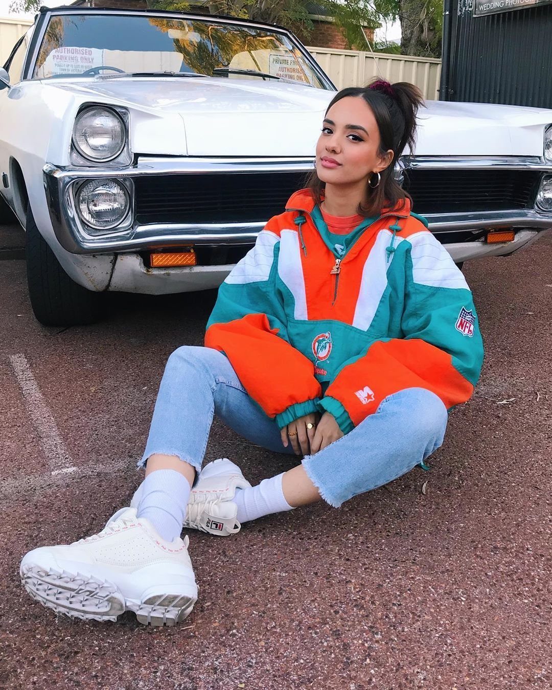 Young woman wearing a colorful windbreaker and denim capris sitting in front of a white car.