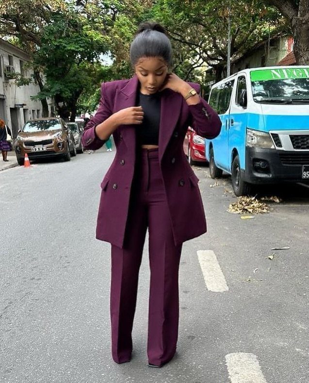 A woman posing in a maroon tailored pantsuit on a city street with trees and vehicles.