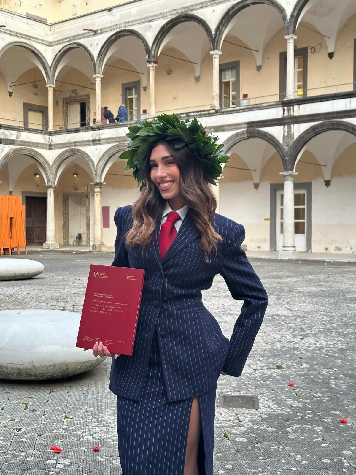 A woman in a navy pinstripe suit and red tie wearing a laurel wreath while holding a red folder.