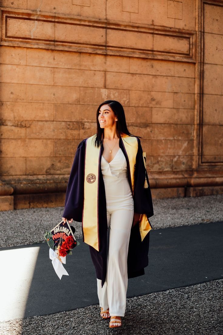 Woman in a white satin wide-leg jumpsuit and graduation gown walking with flowers.