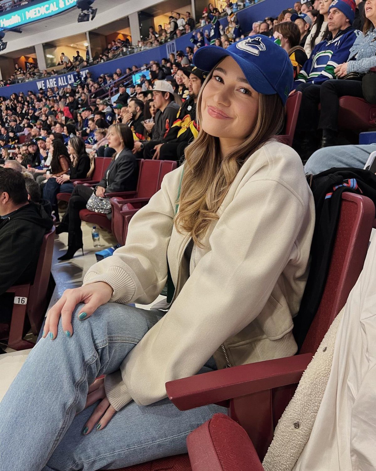 A young woman in a cream jacket and blue cap sitting in stadium seating.