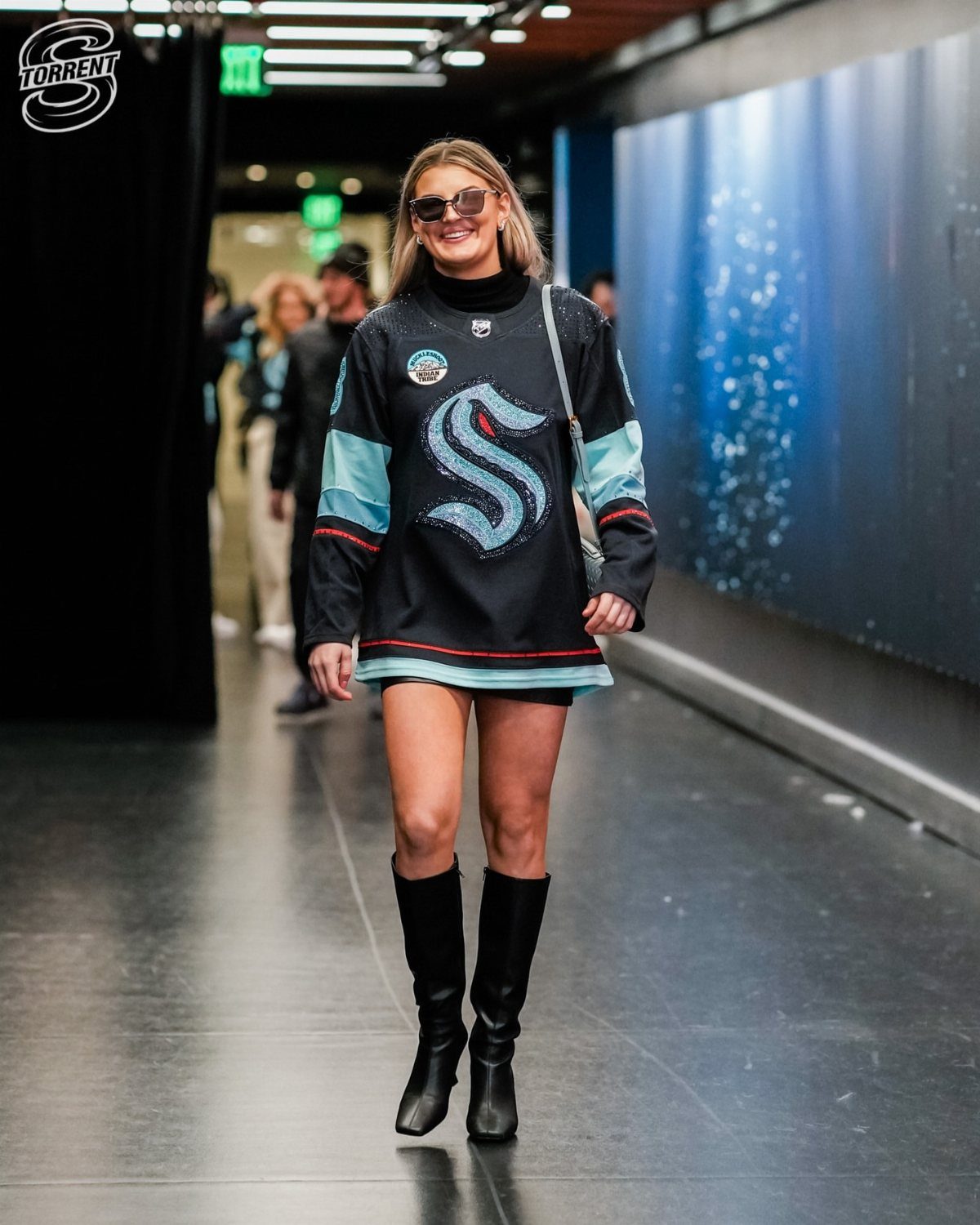 A woman wearing a black hockey jersey as a dress with black boots walking in a hallway.