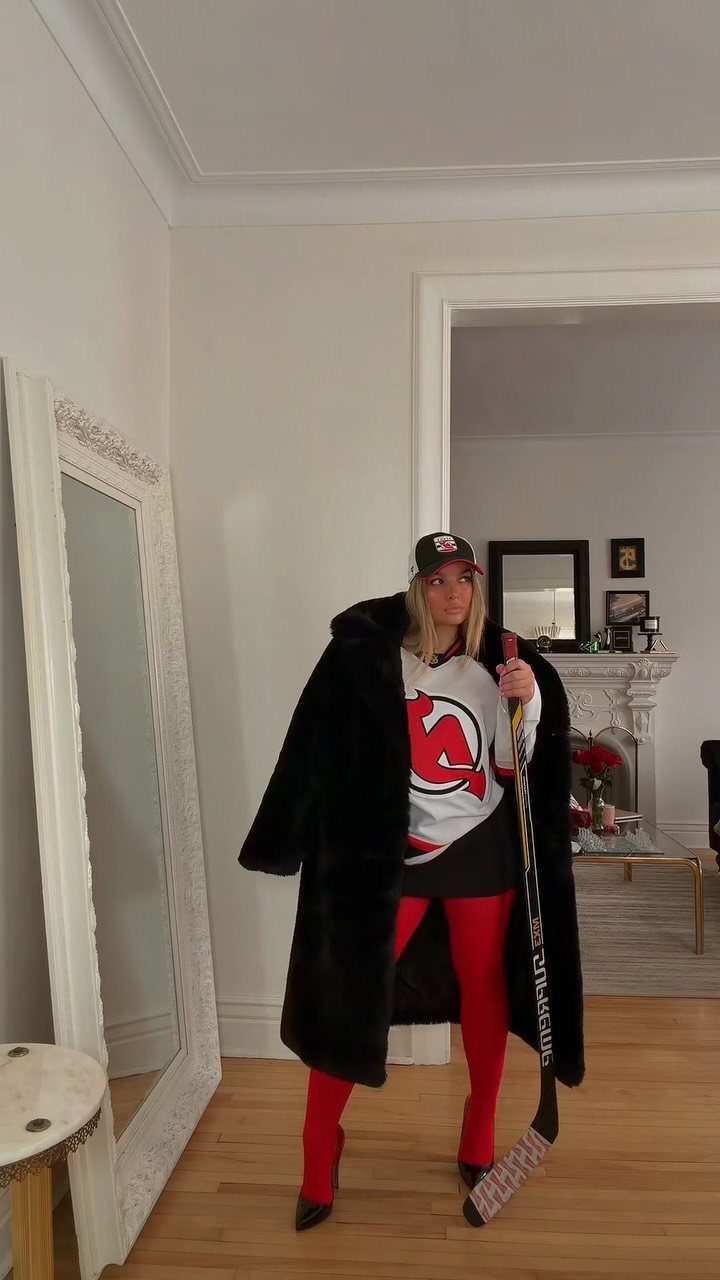 A woman posing in a hockey jersey, black fur coat, and red tights holding a hockey stick.