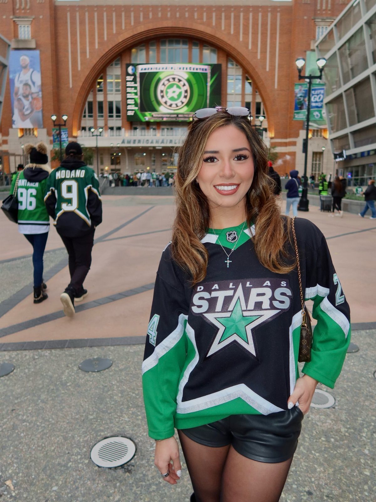 A woman smiling in a Dallas Stars hockey jersey and black leather shorts outside an arena.