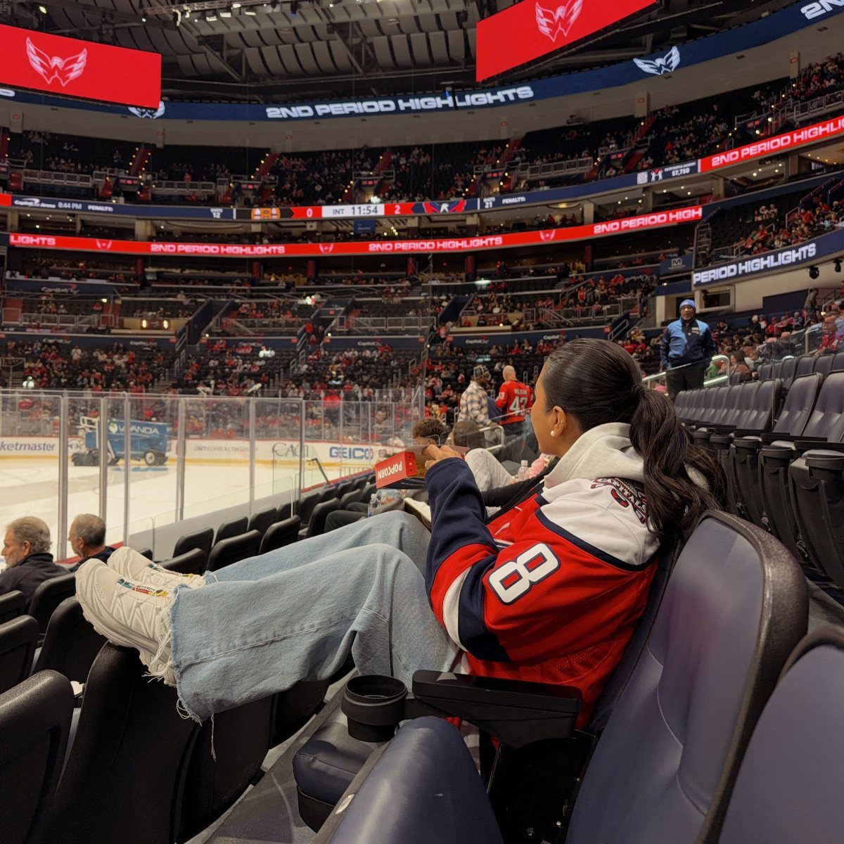 A person wearing a red hockey jersey and light blue jeans sitting in arena seats.
