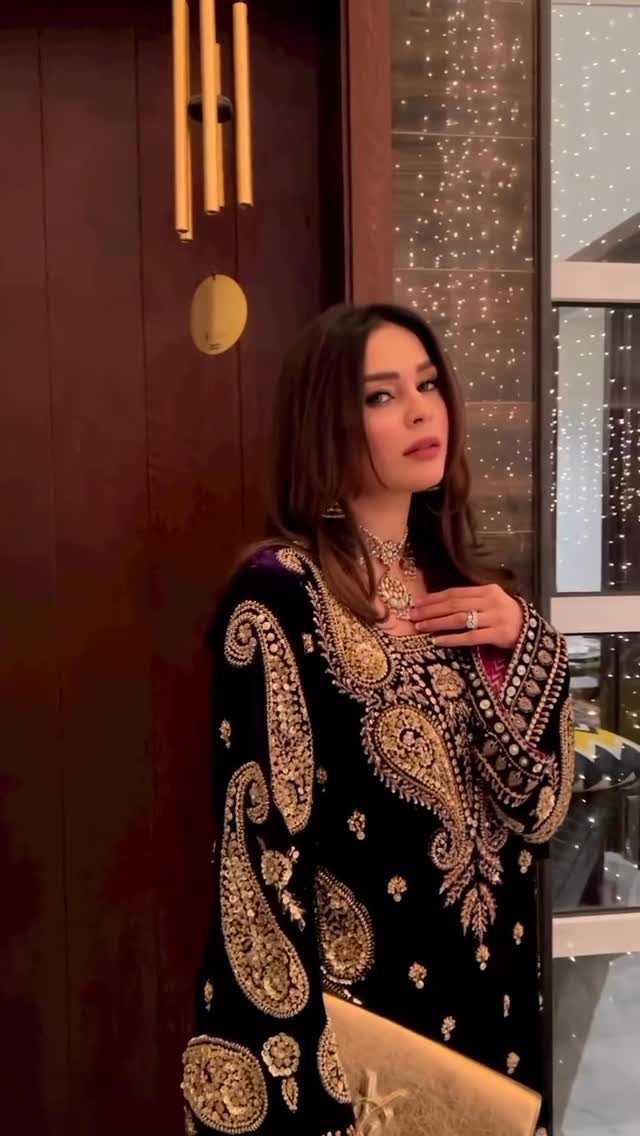 Woman in an ornate black and gold embroidered velvet tunic posing with a gold clutch.