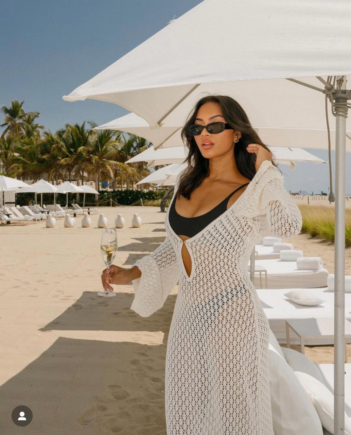 A woman in black swimwear and a white crochet cover-up holds champagne on a sunny beach.