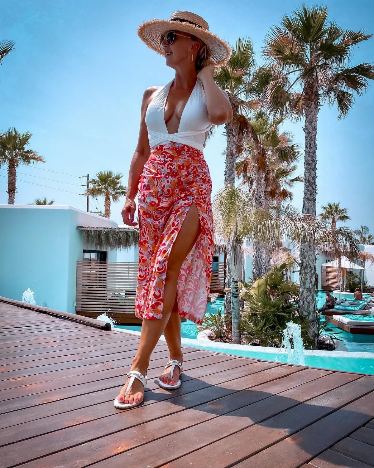 Woman in a white top and patterned skirt walking on a wooden deck wearing white flat sandals.