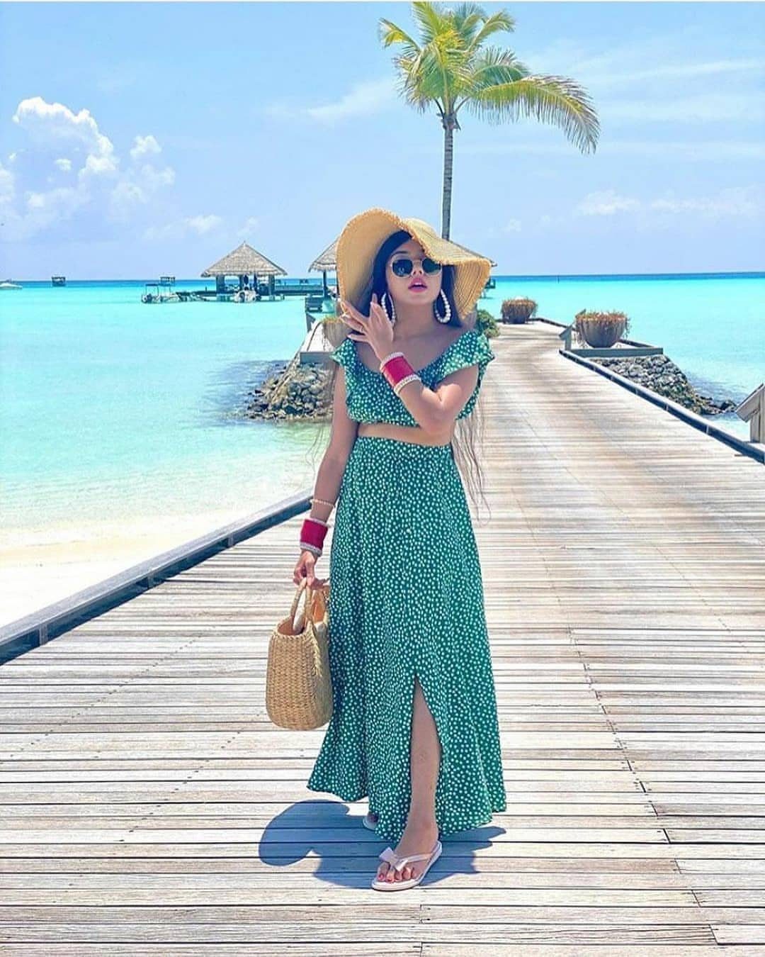 Woman in a green polka-dot outfit and straw hat standing on a wooden pier by the ocean.