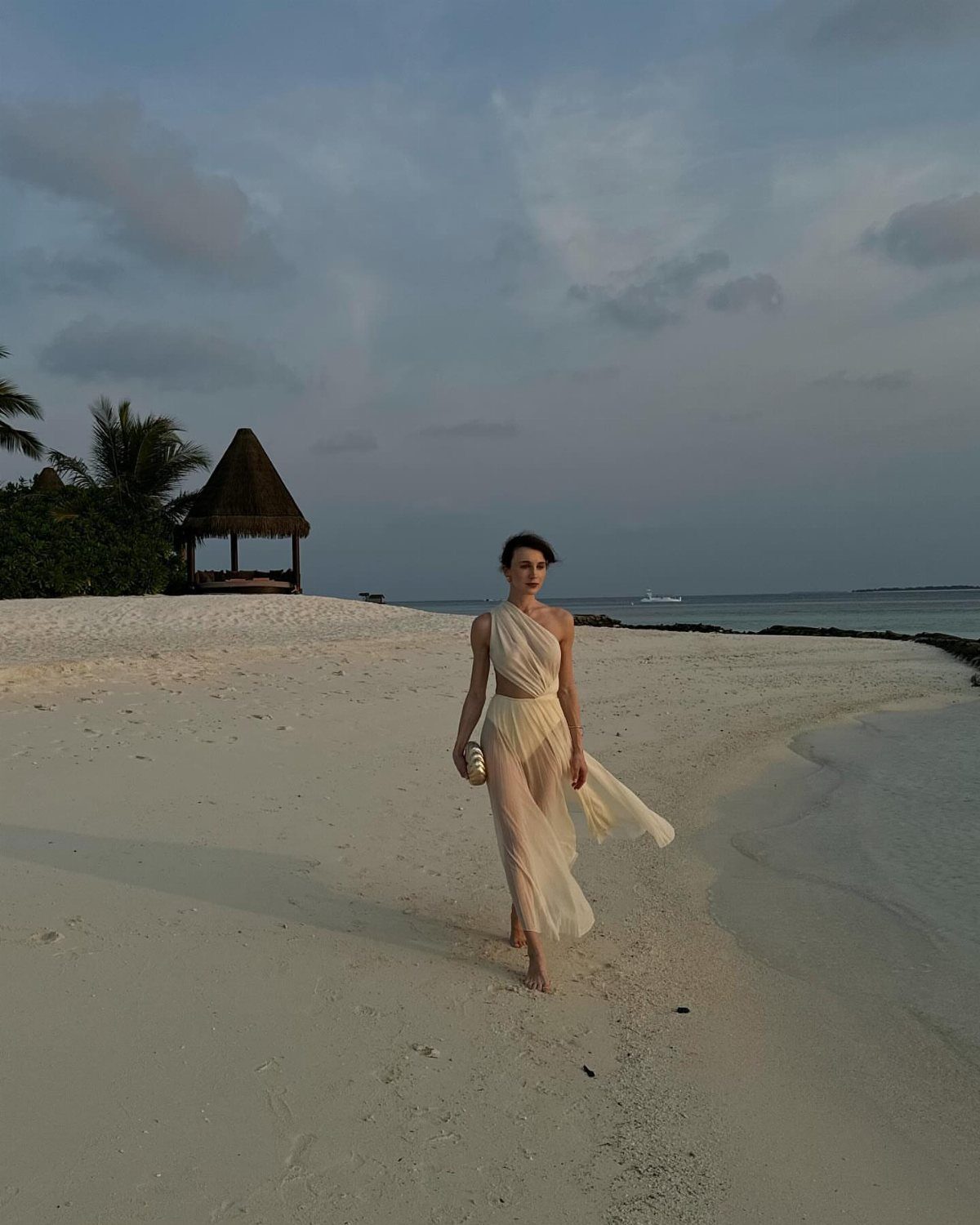 Woman in a flowing white one-shoulder dress walking on a sandy beach at dusk.