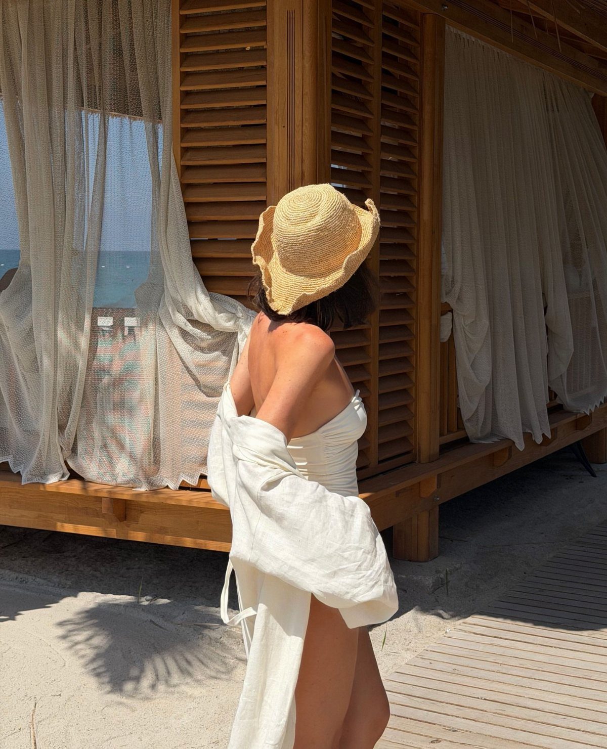 A woman in a white linen outfit wearing a large straw wide-brimmed hat on a sunny beach.