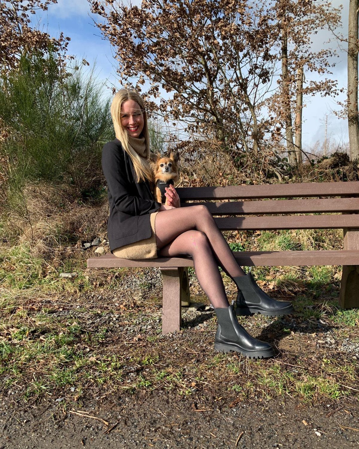 Blonde woman in a black jacket and tan skirt sitting on a bench wearing black Chelsea boots.