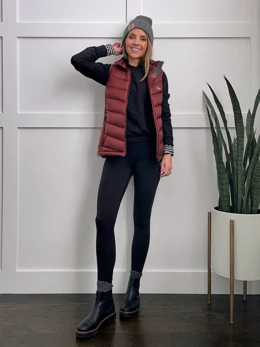 A woman wearing a maroon puffer vest, black leggings, and black Chelsea boots standing indoors.