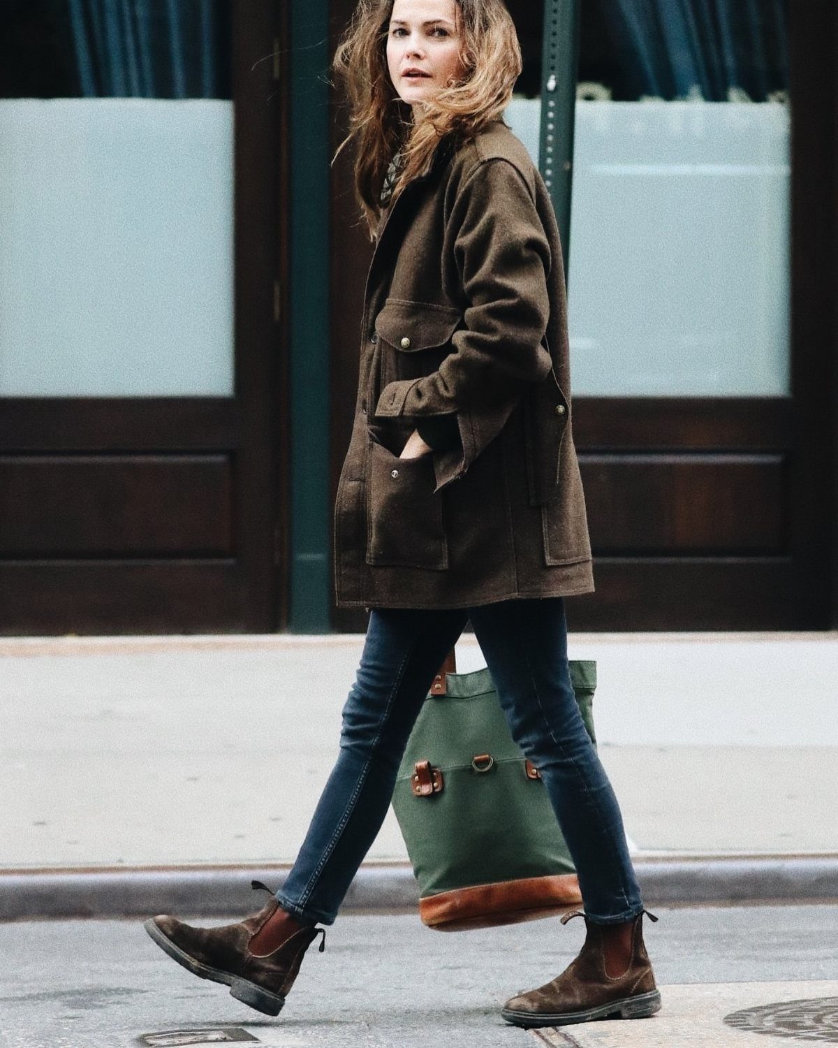 A woman walking in a city wearing a brown jacket, blue jeans, and brown Chelsea boots.