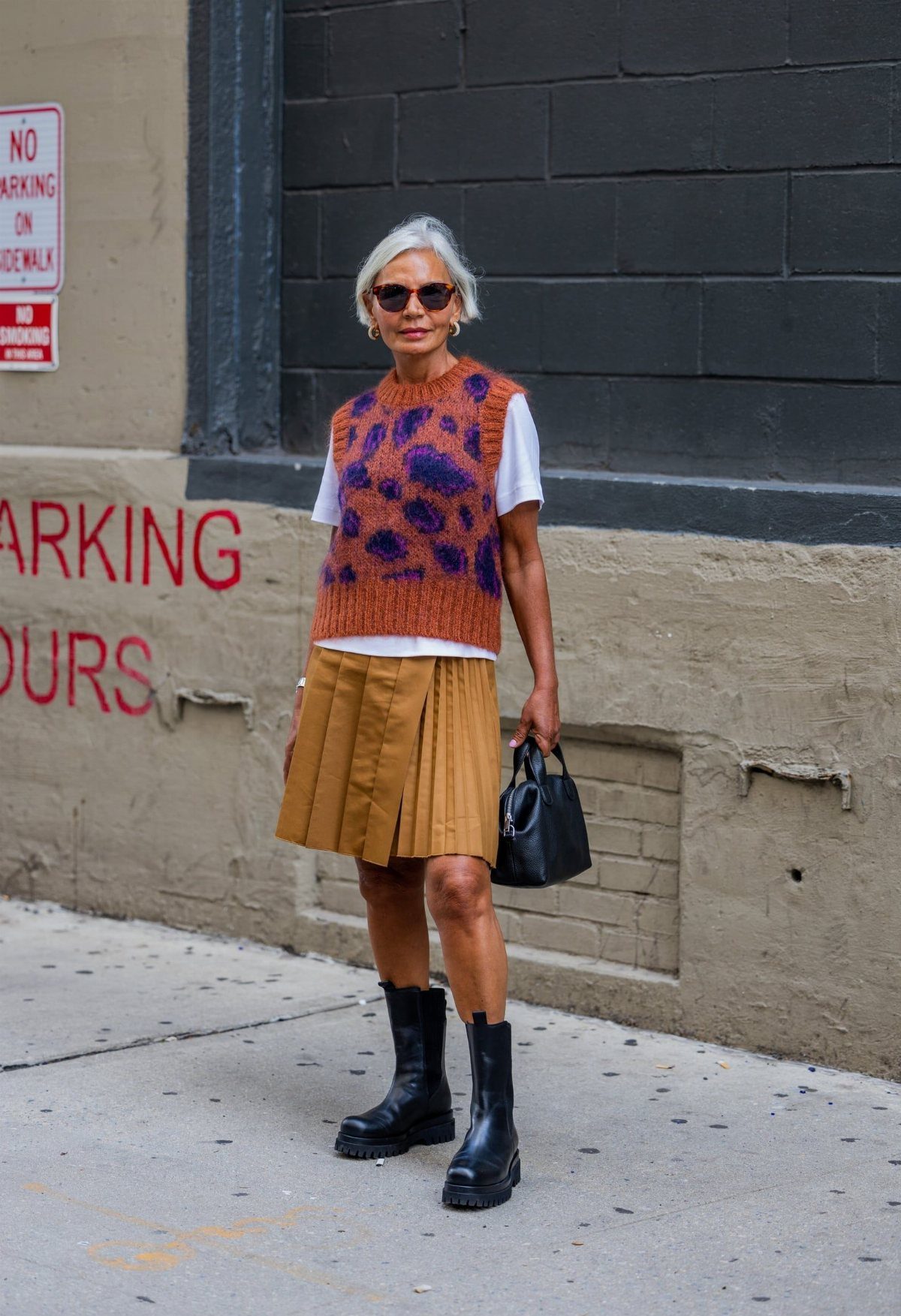 A woman wearing a tan pleated skirt and black Chelsea boots standing on a city sidewalk.