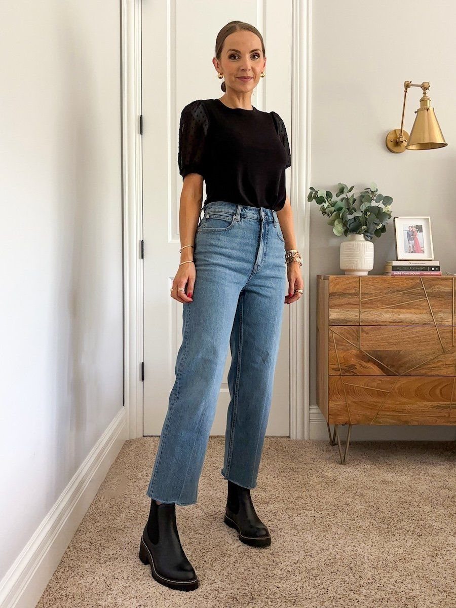 A woman posing in a black top, wide-leg blue jeans, and black Chelsea boots.