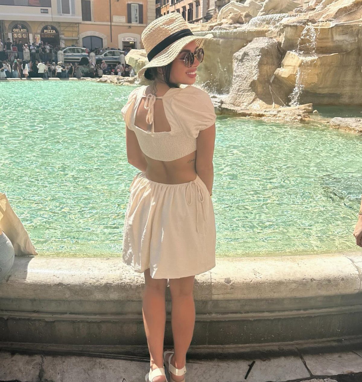 Woman in cream crop top and skirt standing by a turquoise fountain wearing a straw hat.
