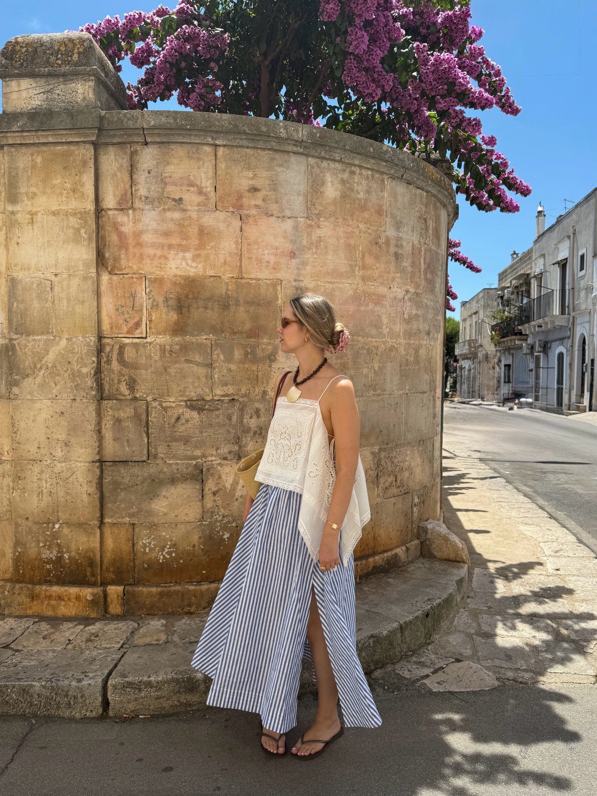 Woman in a white lace sleeveless top and striped maxi skirt standing by a stone wall.
