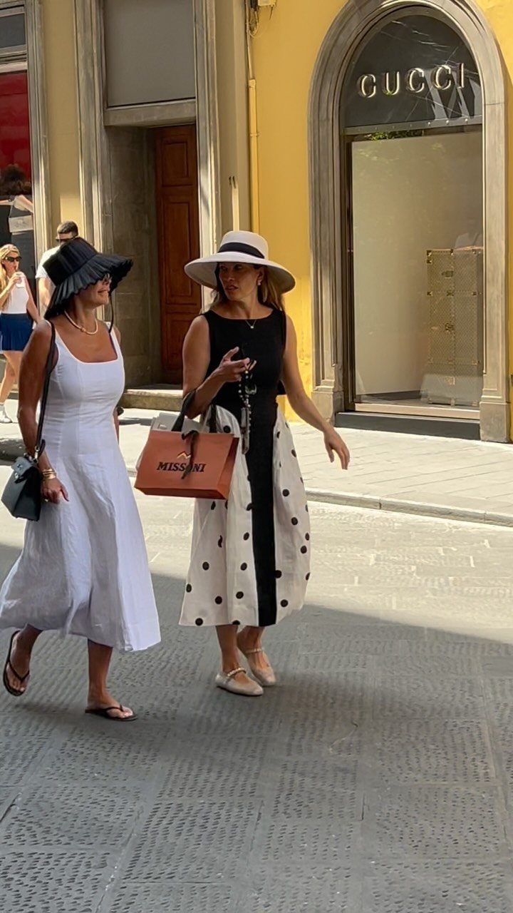 Two women wearing summer sundresses and sun hats walking down a sunny Italian street.