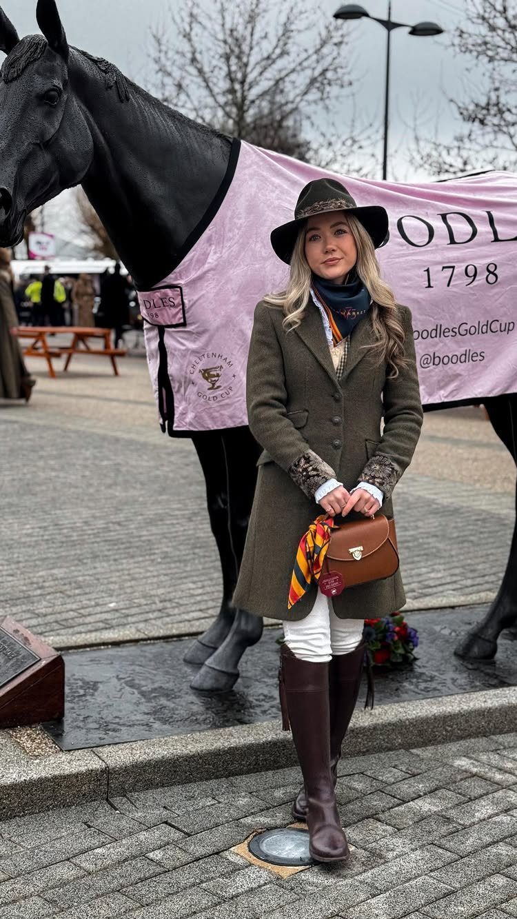 Woman in olive coat, white trousers, and brown riding boots standing by a horse statue.