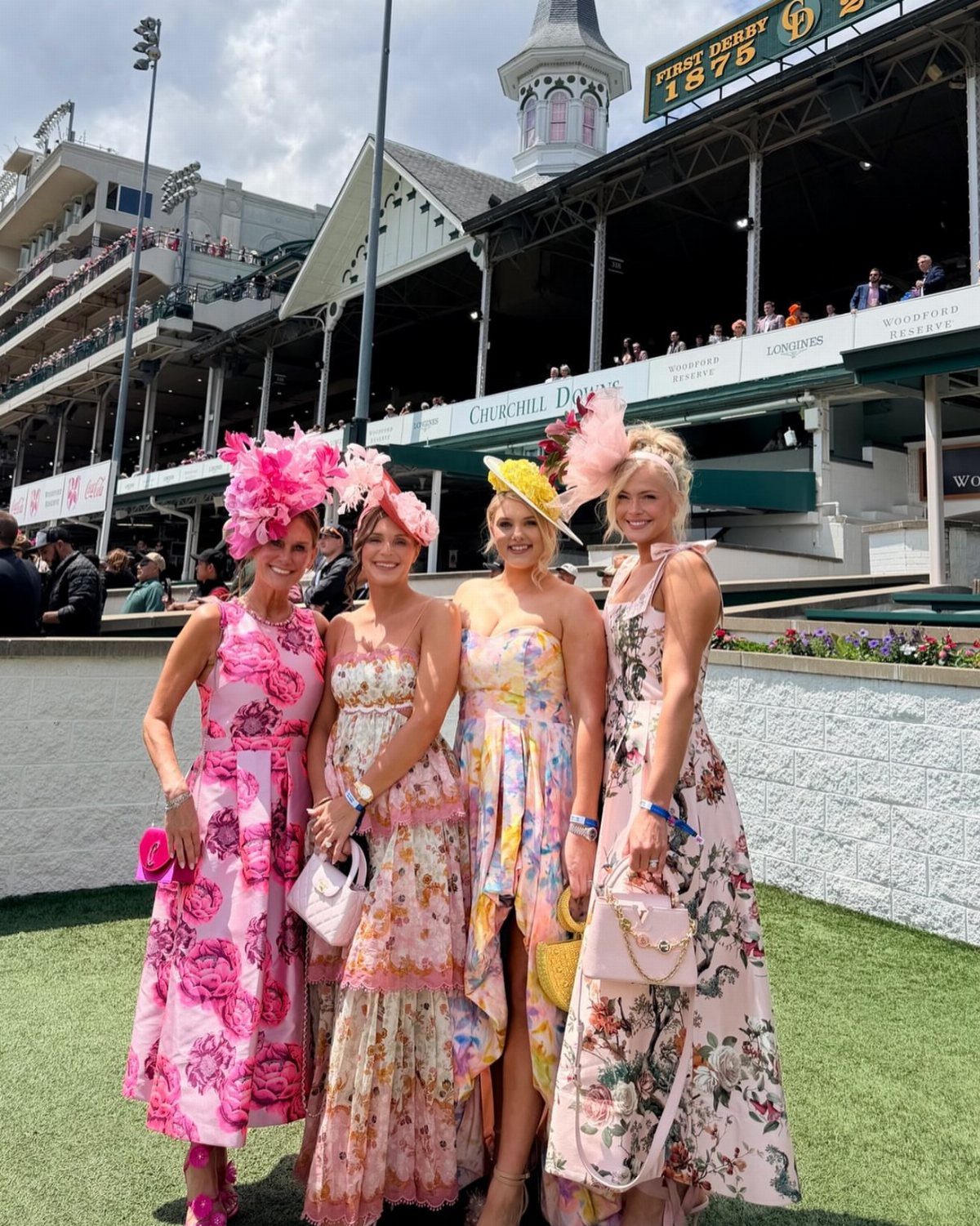 Four women in colorful floral dresses and large decorative hats posing at a racetrack.
