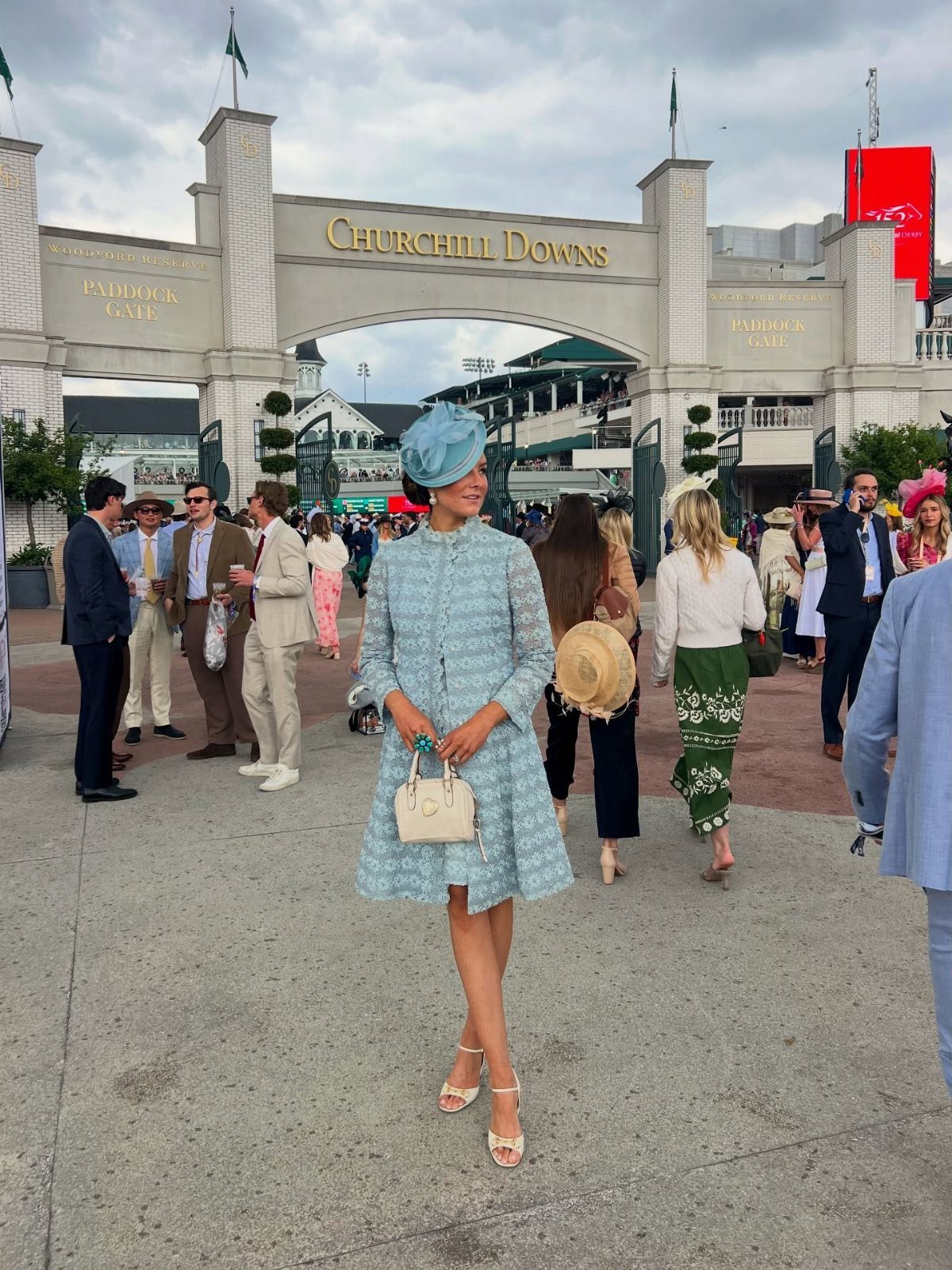 A woman in a light blue dress and fascinator stands before the Churchill Downs gate.