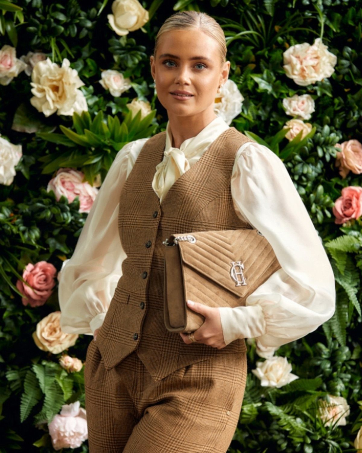 A woman wearing a brown plaid waistcoat and cream blouse posing against a floral wall.