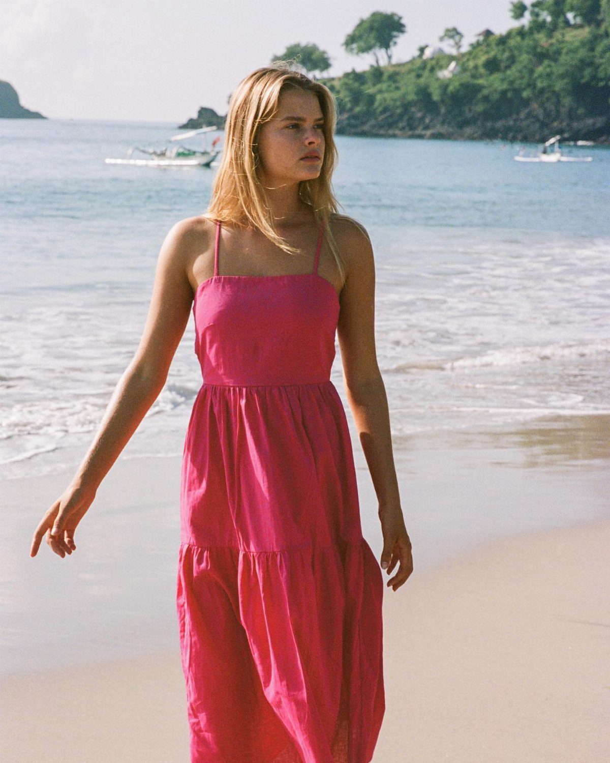 A woman in a bright pink flowy midi dress standing on a tropical beach.