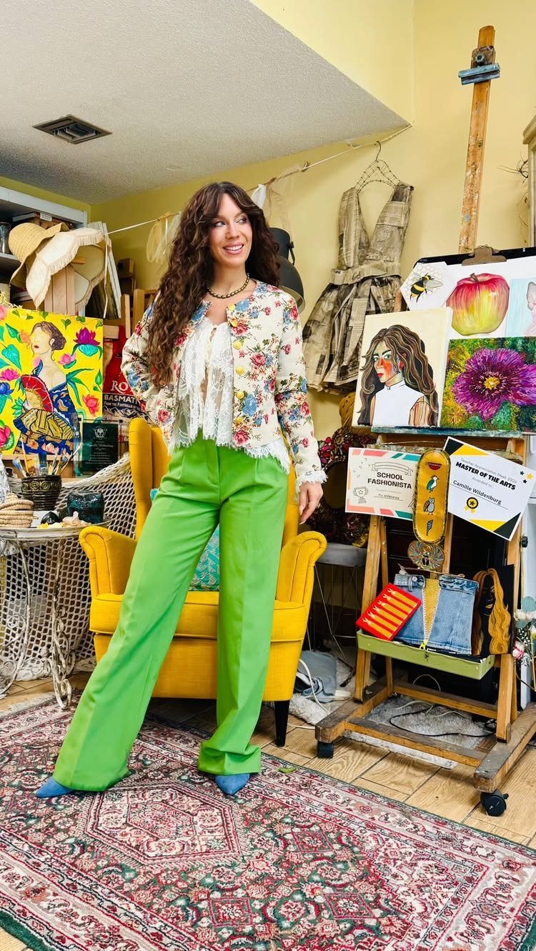 Woman posing in a floral jacket, white ruffled top, and bright green trousers in an art studio.
