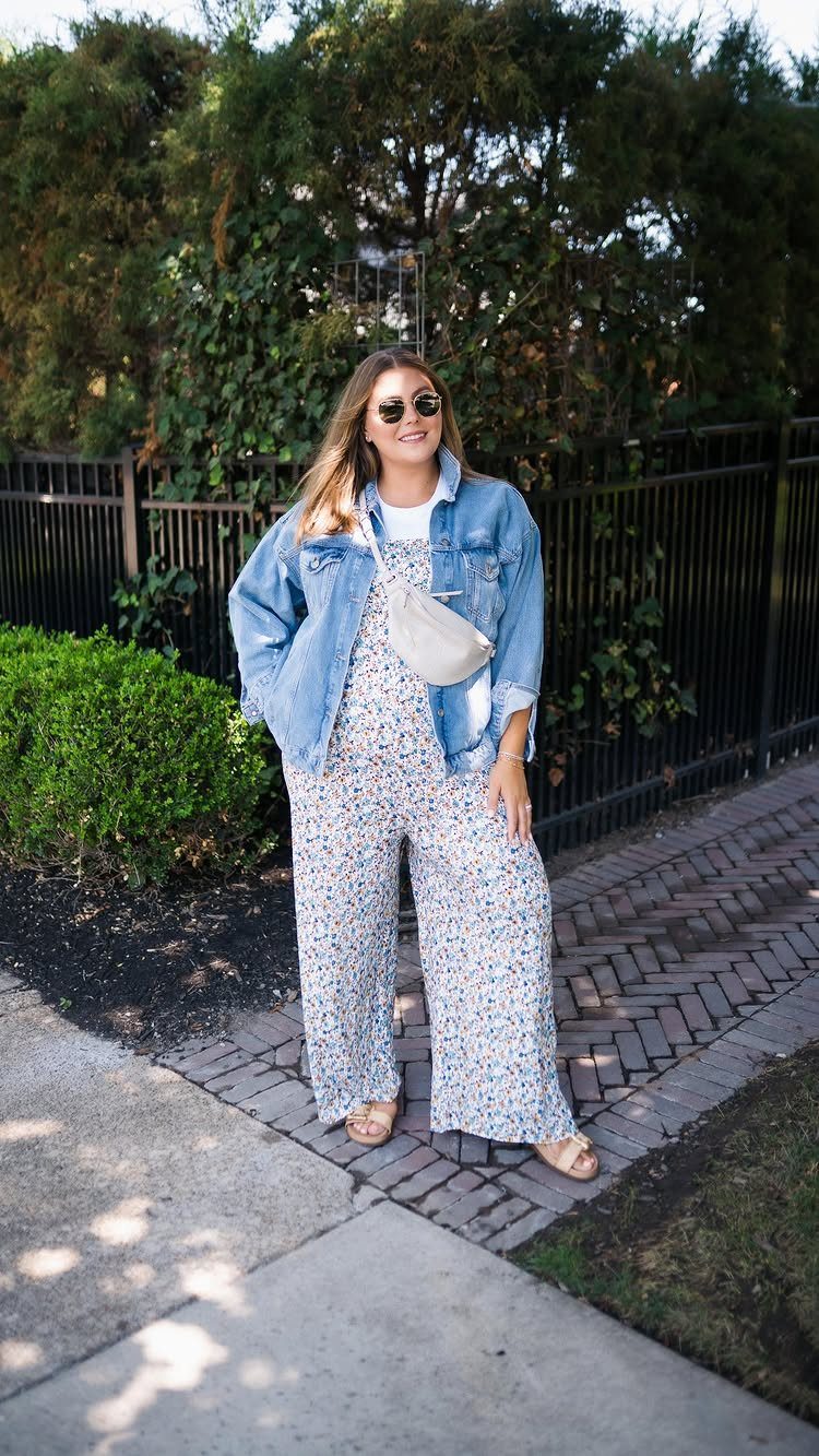 Woman in a floral print jumpsuit and denim jacket standing by a black fence.