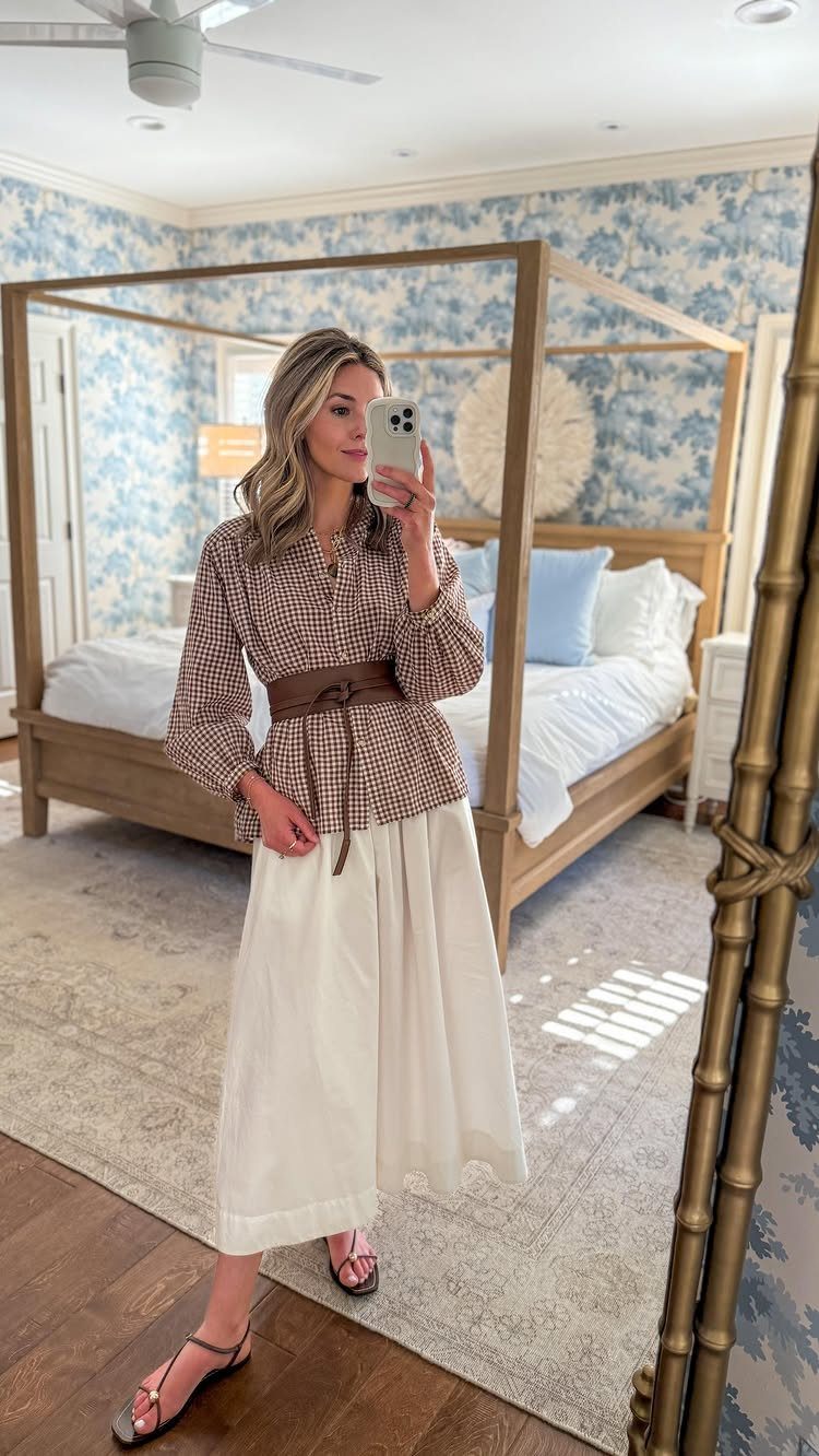 Woman in a patterned blouse, white skirt, and sandals posing for a mirror selfie.
