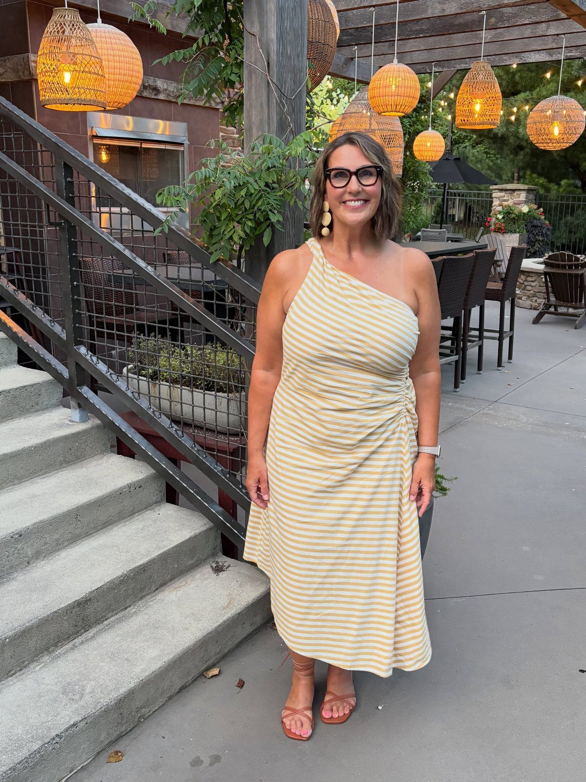 A woman in a yellow striped one-shoulder midi dress standing on a patio.