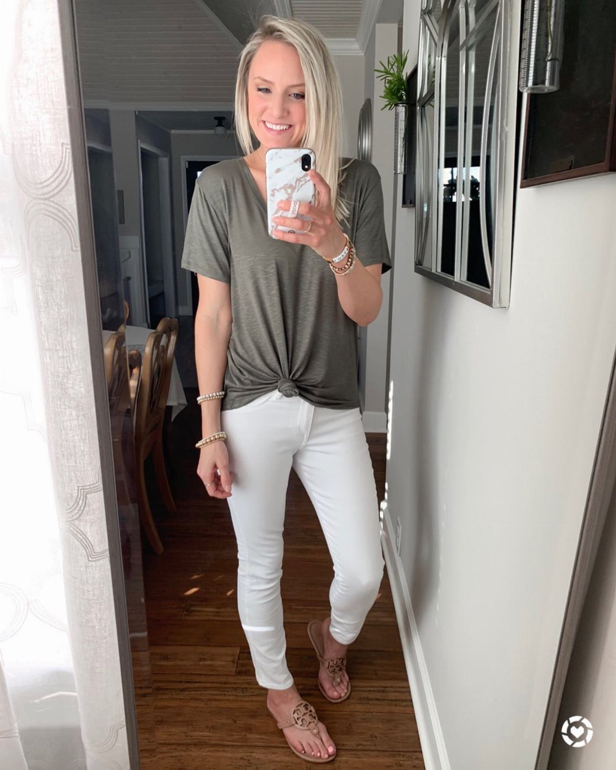 Blonde woman wearing an olive green t-shirt, white pants, and brown sandals standing in a hallway.