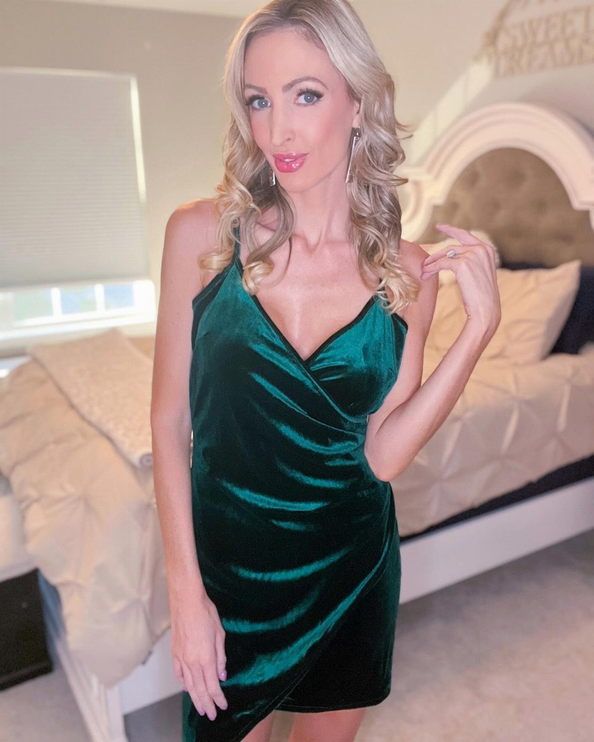 A blonde woman wearing a dark green velvet wrap cocktail dress in a bedroom.