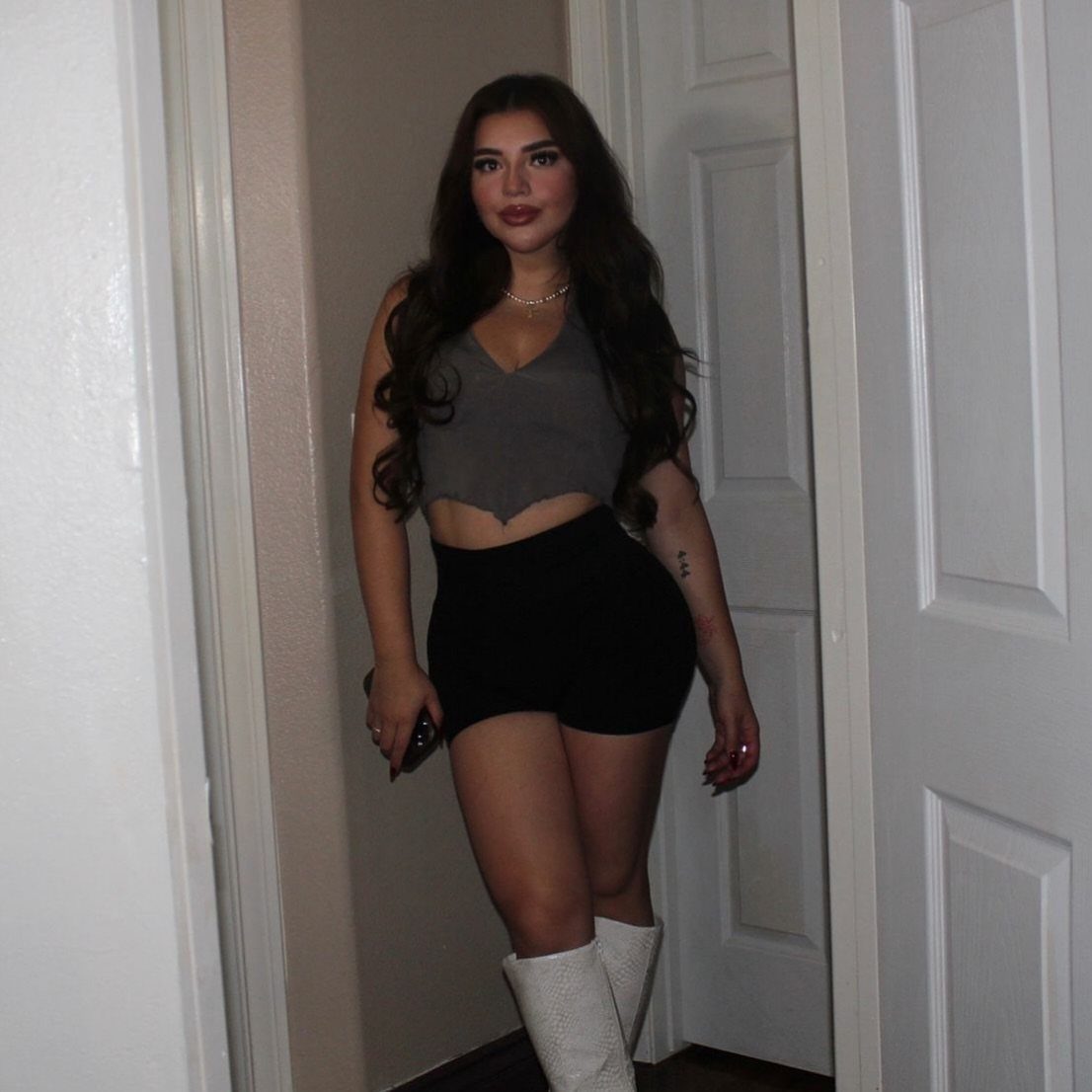 Woman in a grey crop top, black shorts, and white boots posing in a doorway.